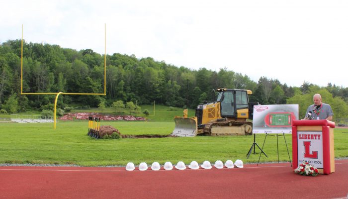A man speaks at a podium with Liberty and a block L with a board featuring the design of the new field and track to his right as well as a bulldozer on the field and a line of white hard hats on the track.