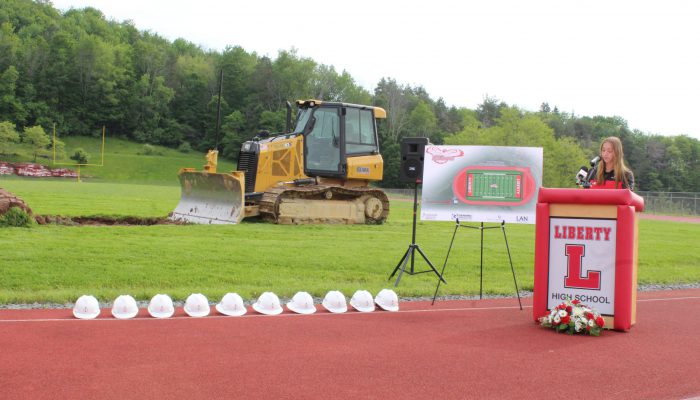 A student speaks at a podium with Liberty and a block L with a board featuring the design of the new field and track to her right as well as a bulldozer on the field and a line of white hard hats on the track.