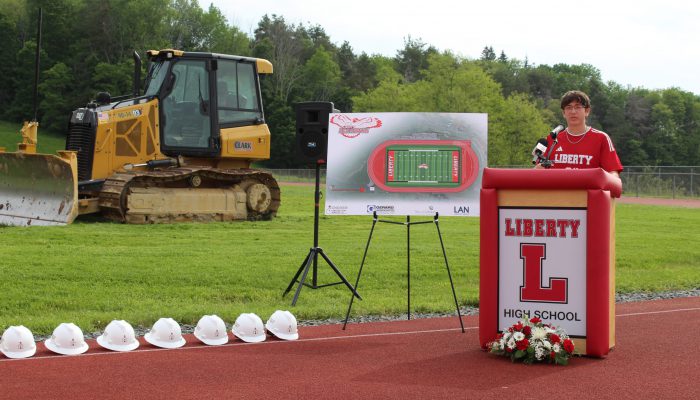 A student speaks at a podium with Liberty and a block L with a board featuring the design of the new field and track to his right as well as a bulldozer on the field and a line of white hard hats on the track.