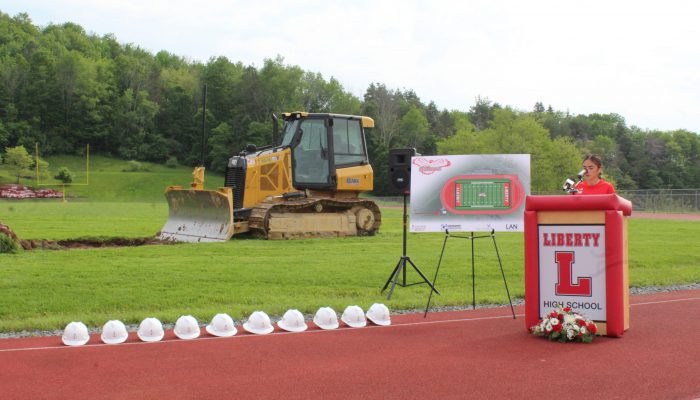 A student speaks at a podium with Liberty and a block L with a board featuring the design of the new field and track to his right as well as a bulldozer on the field and a line of white hard hats on the track.