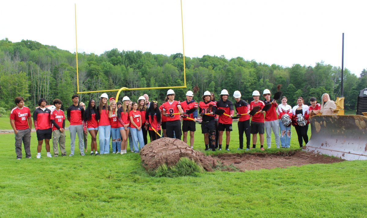 Students wearing hard hats pick up dirt with shovels at a groundbreaking ceremony.