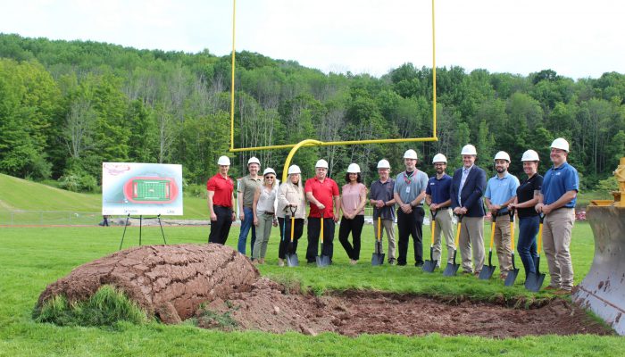 Administrators and officials pose with hard hats next to a pile of dirt and grass with a board featuring the new field and track next to them.