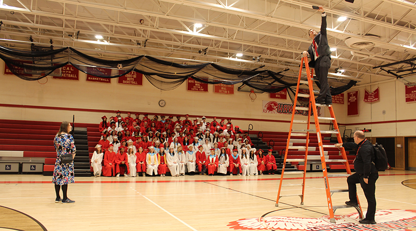 A man takes a selfie with standing near the top of a ladder with graduates seated in bleachers behind them as another man holds the ladder and a woman watches at left