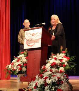 A woman speaks at a podium with the Liberty Readhawks logo as a man stands near.