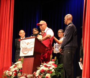 A man stands at a podium with the Liberty Redhawks logo on it surrounded by family. and friends.