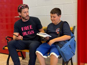 An adult sits with "free hugs" on his shirt sit next to a student as he hands him a book