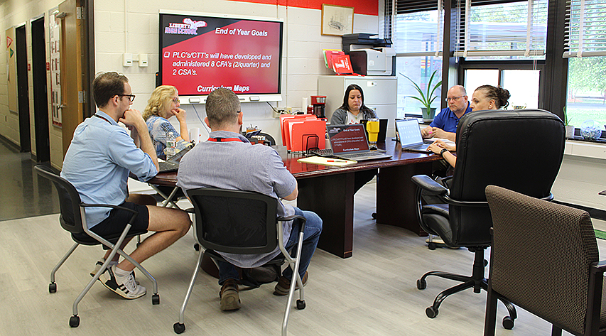 People sit around a table and talk with a large screen behind listing an end of year goal them