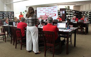 People sit or stand around tables set up in a U shape as a woman speaks at back.
