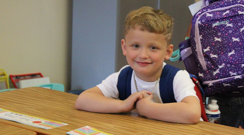 A student wearing a backpack smiles while seated at a table.