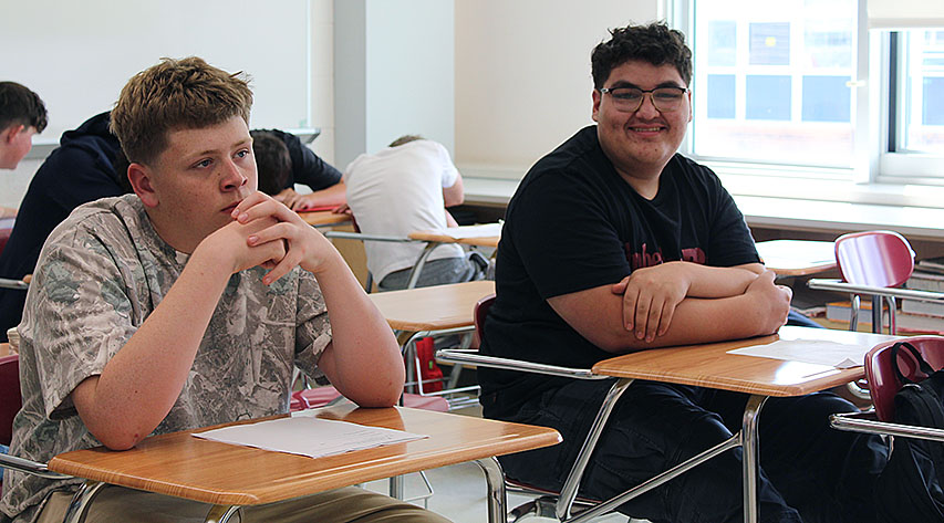 A student smiles seated at a desk while a another student looks ahead