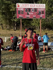 A soccer player holds up four fingers and poses with the man of the match medal in front of the scoreboard.