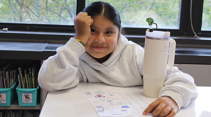 A student leans on her hand and smiles as she works on a worksheet with a large white water bottle next to her