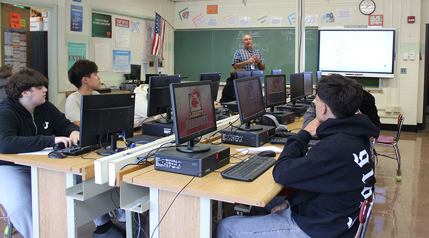 Students listen to a teacher speak while seated at tables with desktop computers on them