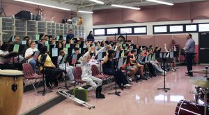 Students seated in chairs on risers with music stands in front of them raise their hands as an adult speaks at the front of the room