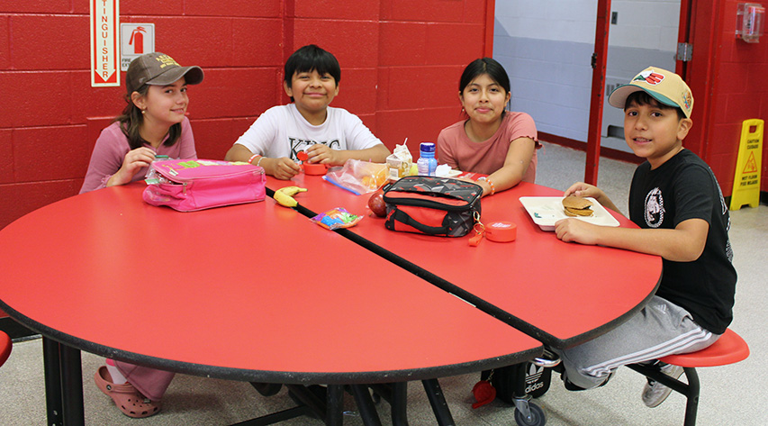Four students smile as they sit at a circular table while eating lunch