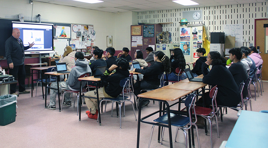 A teacher leads a lesson in front of students seated at lines of desks.