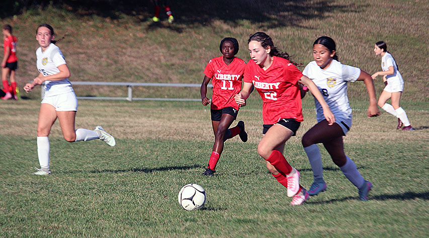 A girls soccer game as a girl in red approaches a ball and a girl in white comes up from behind as other teammates run near.