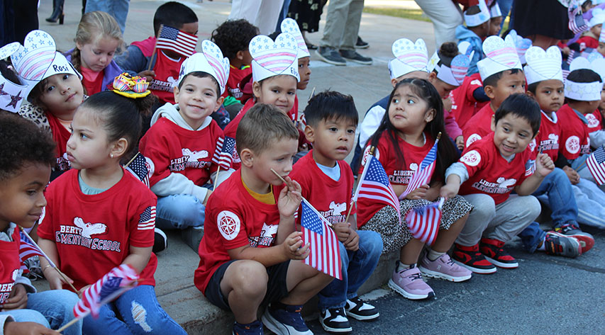 Students in red shirts and some wearing red white and blue paper hats hold American flags as they sit on a curb and sidewalk.