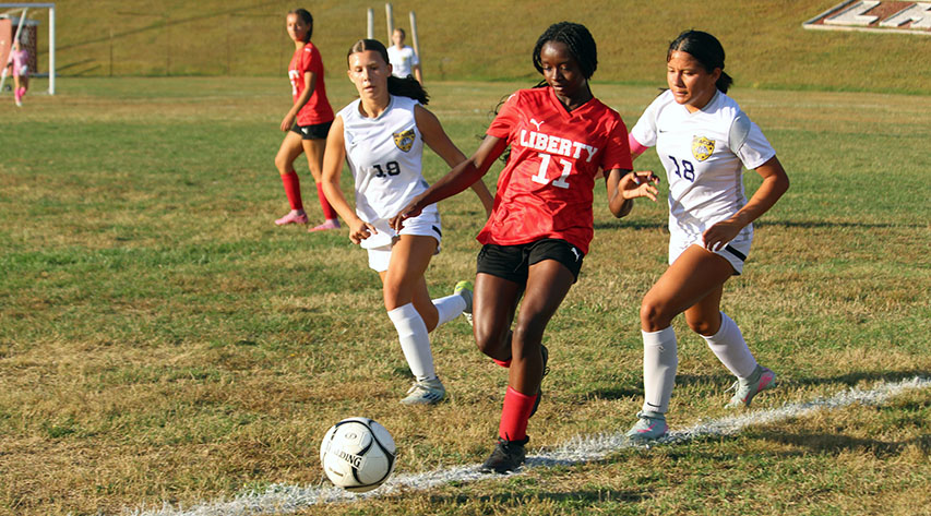 A soccer player in red approaches the ball as two players in white come up behind her