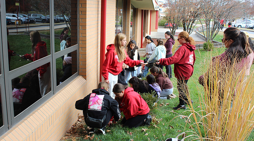 A student holds another student a tools as students and family work to plant a daffodil border in front of a school