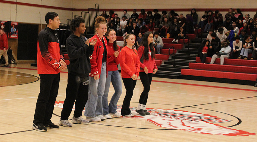 Six students stand on the center line near the logo in the gym