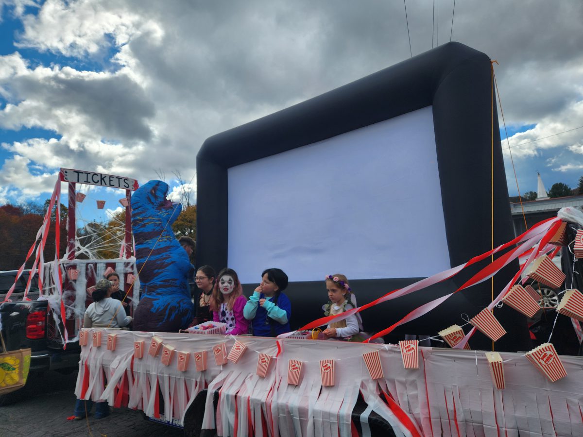 A truck with a ticket booth in the back tows a trailer with an inflatable movie screen with students dressed in costumes