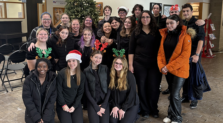 Students pose in front of a decorated tree, some with Santa hats on
