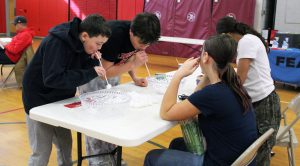 Two students race to move cotton balls from a pile to bowls using straws.