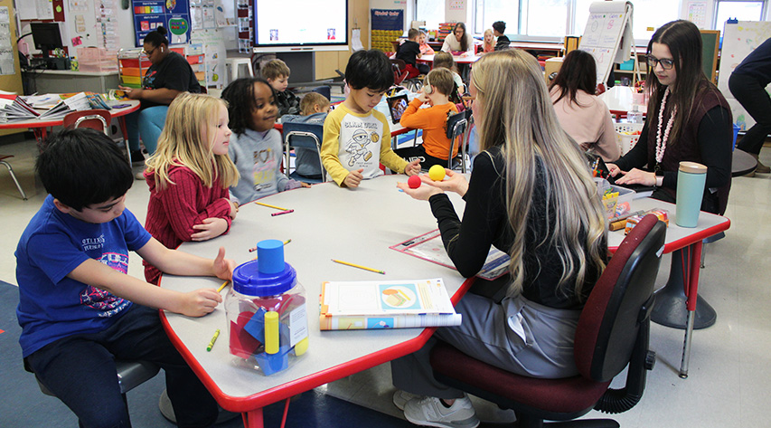 A teacher displays two balls as students sit around a table as another teacher observes and other students and teachers work at tables in a classroom.