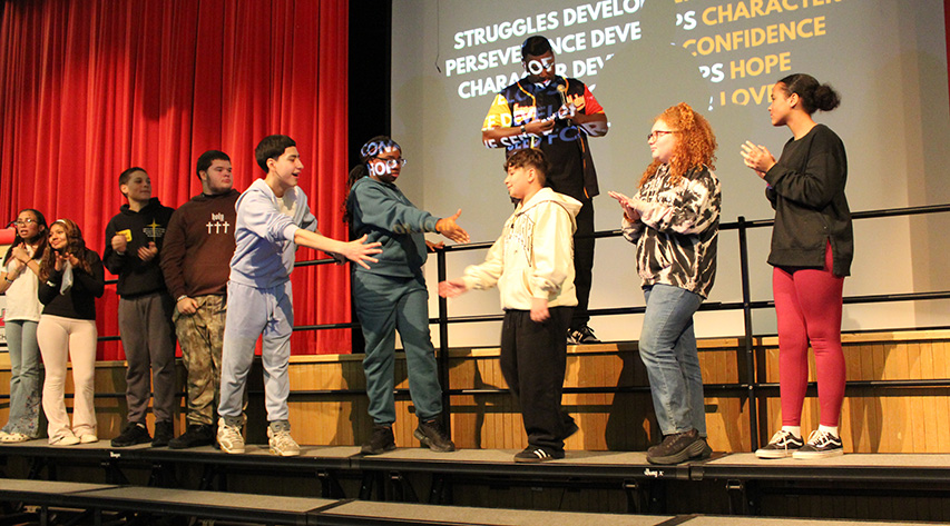 Two students reach out to shake the hand of another student as other students stand on risers and adult stands behind him with a projector on him.