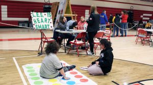 Two students play twister in front of a sign that says LES is Heart Smart next to a table with three adults around it.