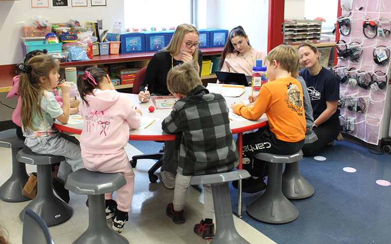 Teachers observe a lesson as another teacher works with students around a table.