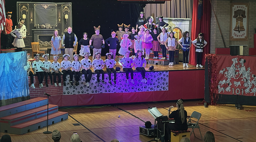 Students stand on stage as the director sits a front during a production of 101 dalmatians kids.