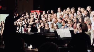 A director in shadow leads a choir as a pianist plays