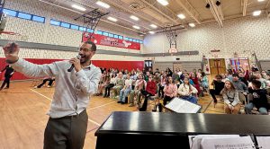 A man takes a selfie while standing in front of student seated in Liberty Middle School gym.