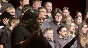 A student plays the flute as others sing behind her.