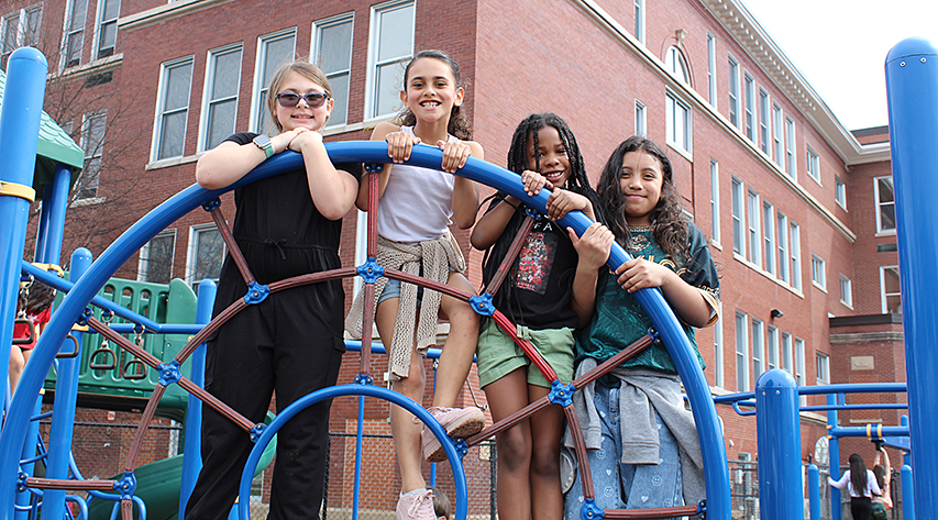 Four students smile while posing for a photo on a piece of playground equipment