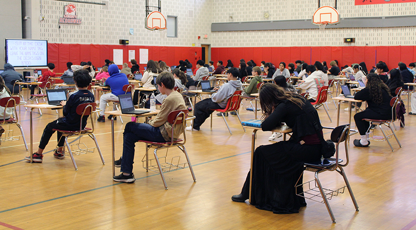 Students sit at desks taking tests on computers in rows