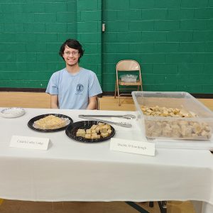 A student smiles as while sitting behind a table with food.