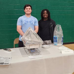 Two students pose for a photo behind a buffet tray and plates on a table