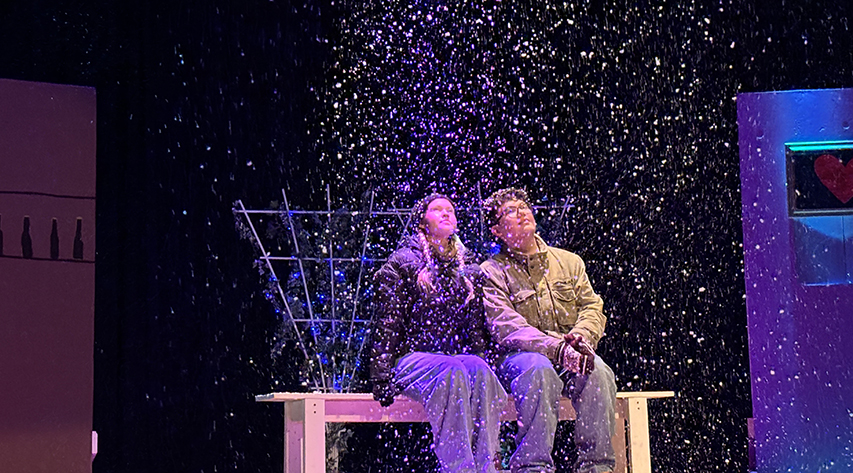 Two students sitting on a bench look up as it snows on a stage