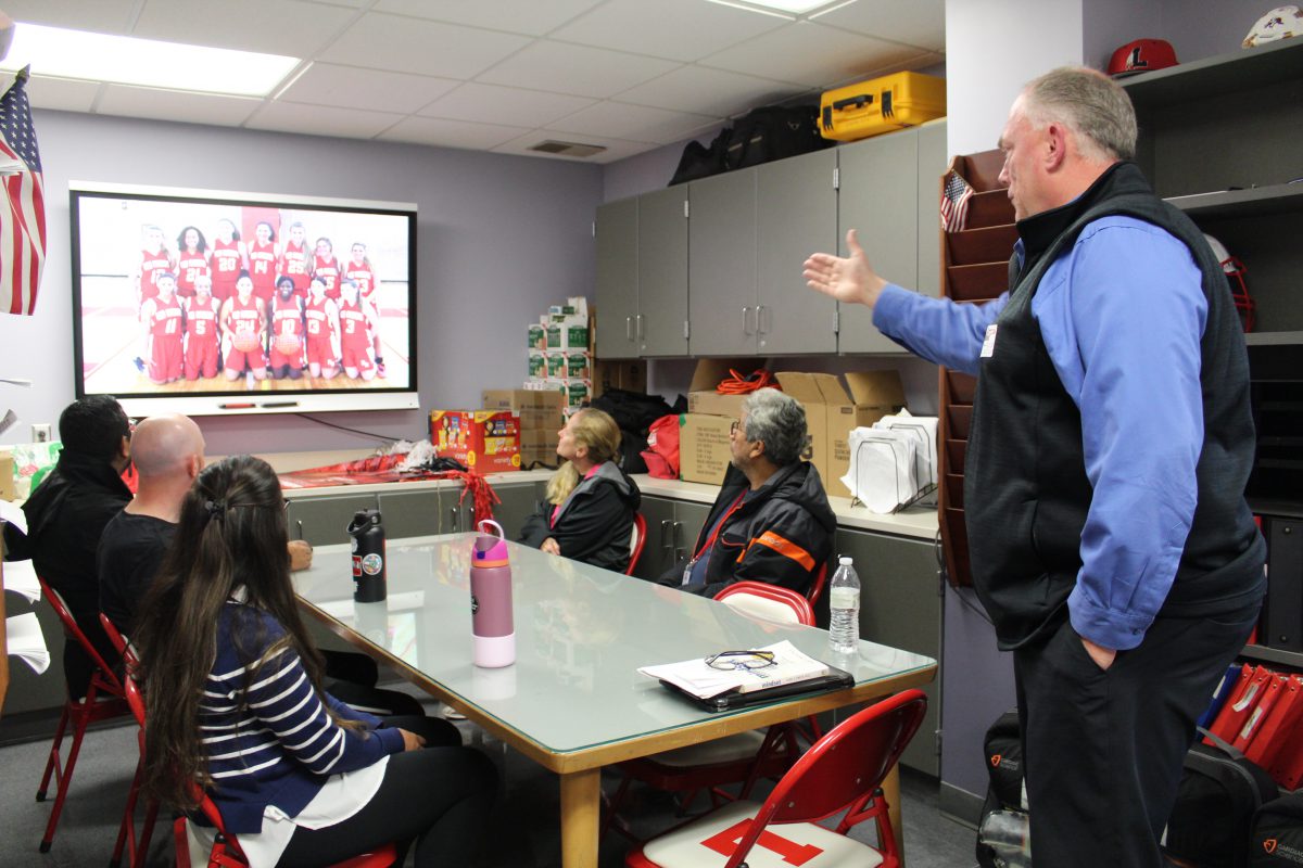 A man gestures to a screen featuring a picture of a girls basketball teams as people sitting at a table look.