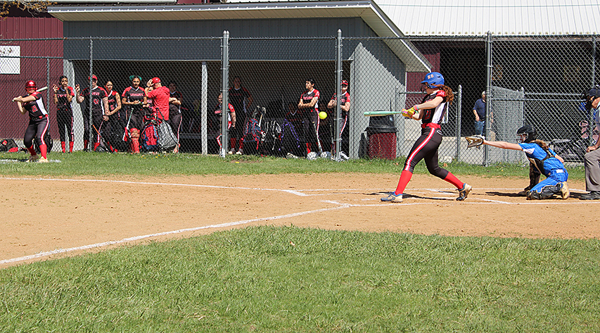 A batter hits a softball as her team watches from the sidelines and the catcher reaches out.