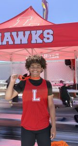 A track athlete holds his medal while standing in front of a Redhawks tent
