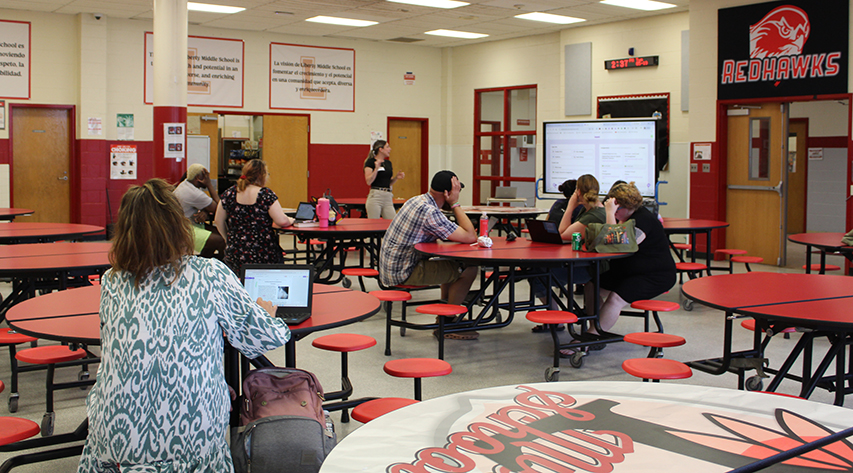 A woman speaks at a smartboard as adults sit at cafeteria tables during a professional development session.