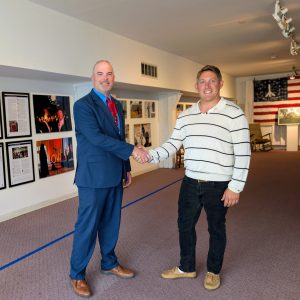 Two men shake hands in a gallery space with framed artwork on the walls.