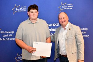 A young man in a grey polo shirt holds a certificate while standing next to a man in a plaid blazer, both smiling in front of a blue backdrop with the Sullivan BOCES logo.