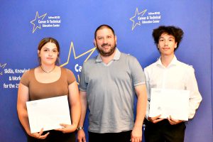 A man stands between two young people, all holding certificates in front of a blue backdrop with stars and text.