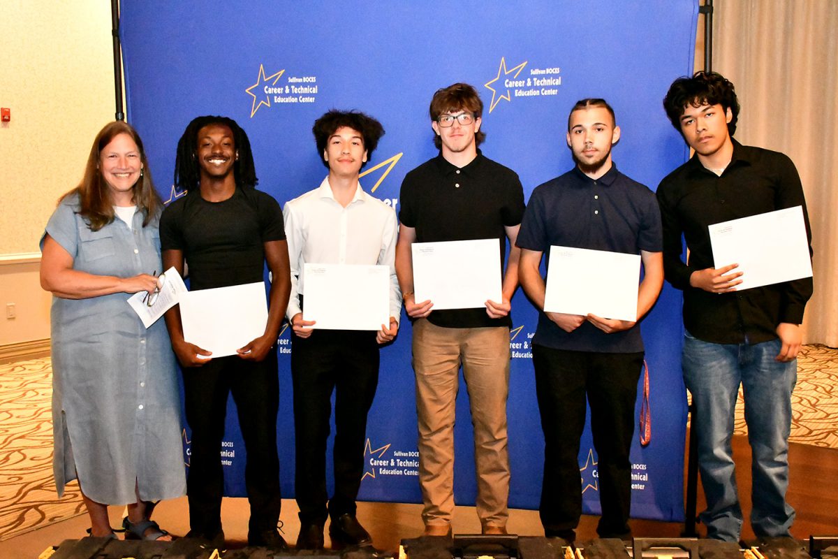 A woman in a blue dress stands with five young men, each holding a certificate, in front of a blue banner.
