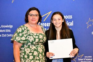 A woman in a floral dress and a young woman in a black jacket smile, holding a certificate in front of a blue backdrop with text.